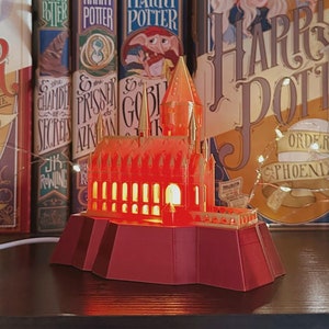 May include: A red and gold Hogwarts castle light-up model with a lit interior. The model is on a shelf with Harry Potter books and figurines.