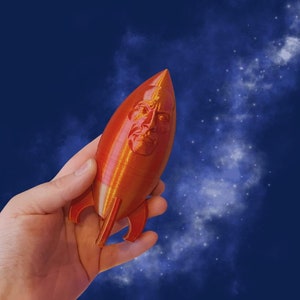 May include: A red rocket-shaped toy with a human face on the front. The toy is being held in a hand against a blue background with white clouds.