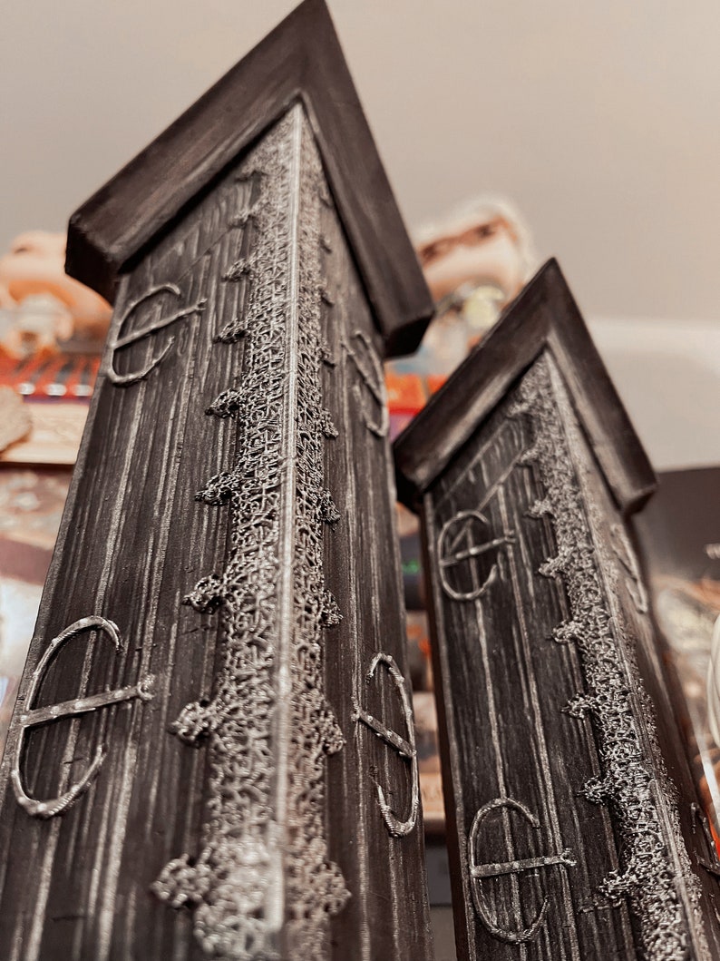 May include: Two tall, black wooden towers with intricate white designs and a pointed top. The towers have a weathered look and are decorated with a repeating pattern of circles.