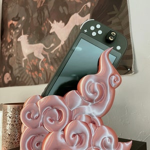 Könnte beinhalten: Ein roségoldener, 3D-gedruckter Wolkenständer für eine Nintendo Switch-Konsole. Der Ständer ist so konzipiert, dass er die Konsole aufrecht hält, wobei der Bildschirm nach außen zeigt.