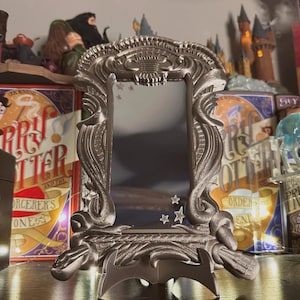 May include: A silver-colored, ornate mirror with a stand. The mirror has a decorative frame with a design of stars and a crest. The mirror is on a shelf with Harry Potter books.