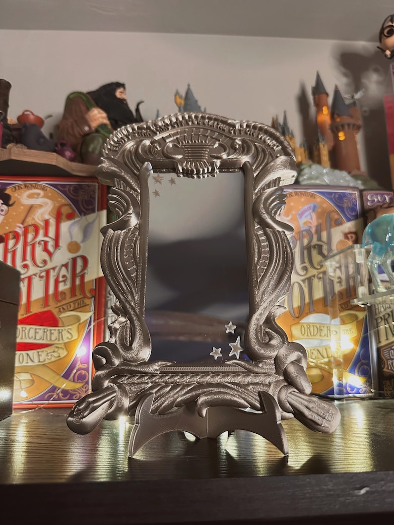 May include: A silver-colored, ornate mirror with a stand. The mirror has a decorative frame with a design of stars and a crest. The mirror is on a shelf with Harry Potter books.