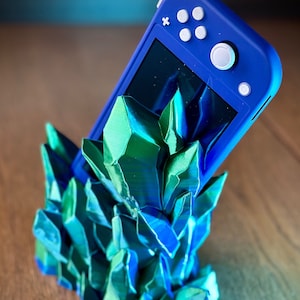 Puede incluir: Una consola Nintendo Switch Lite azul descansa en un soporte impreso en 3D hecho de cristales verdes y azules.