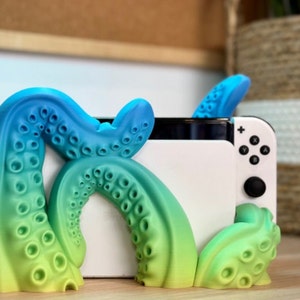 Puede incluir: Un soporte de pulpo impreso en 3D para una consola Nintendo Switch. El soporte es de color verde, azul y blanco y está formado como un pulpo con tentáculos que sujetan la consola en su lugar.