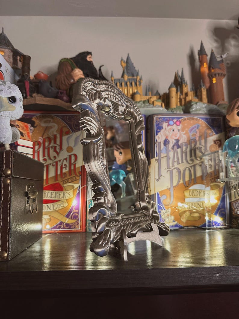 May include: A silver ornate mirror with a decorative frame, sitting on a shelf with Harry Potter themed books and figurines.