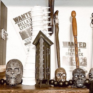 May include: A collection of Halloween decorations including a skull clock, a skull with a snake, a small house, a white candle holder, a black and white mask, and two wands. The decorations are arranged on a shelf with a wooden base.