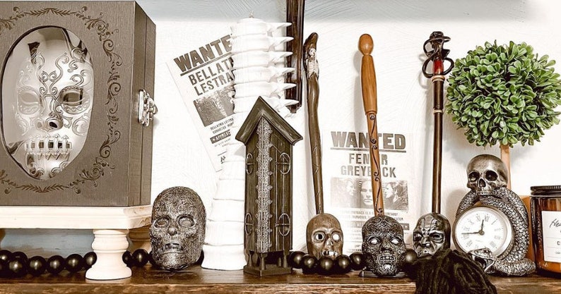 May include: A collection of Halloween decorations including a skull clock, a skull with a snake, a small house, a white candle holder, a black and white mask, and two wands. The decorations are arranged on a shelf with a wooden base.