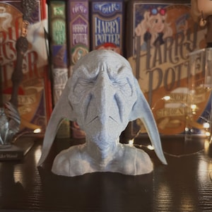 May include: A gray 3D printed bust of a house elf with large ears. The bust is sitting on a black shelf with a collection of Harry Potter books behind it.