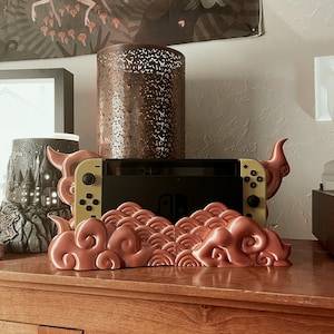 May include: A pink and gold Nintendo Switch console stand with a cloud and wave design. The stand is made of plastic and has a textured finish.
