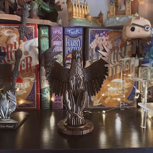 Puede incluir: Una figurita esculpida en marrón oscuro de un fénix con las alas extendidas, de pie sobre una base circular. La figurita está en un estante de madera oscura con una fila de libros de Harry Potter detrás.