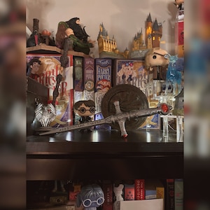 May include: A shelf with a collection of Harry Potter books, figurines, and a sword. The books are in a row, with the titles "Harry Potter and the Prisoner of Azkaban", "Harry Potter and the Goblet of Fire", and "Harry Potter and the Order of the Phoenix". There is a small figurine of Harry Potter in front of the books, and a sword is lying on top of the books.
