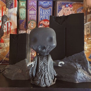 May include: A gray figurine of a character from the Harry Potter series, with a pale face and dark robes. The figurine is posed in front of a black backdrop and gray rock formations, with Harry Potter books in the background.