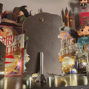 May include: A black metal silhouette of a wizard with a pointed hat and a long beard. The silhouette is on a stand and has a gold accent on the top. The silhouette is in front of a bookshelf with Harry Potter books and figurines.