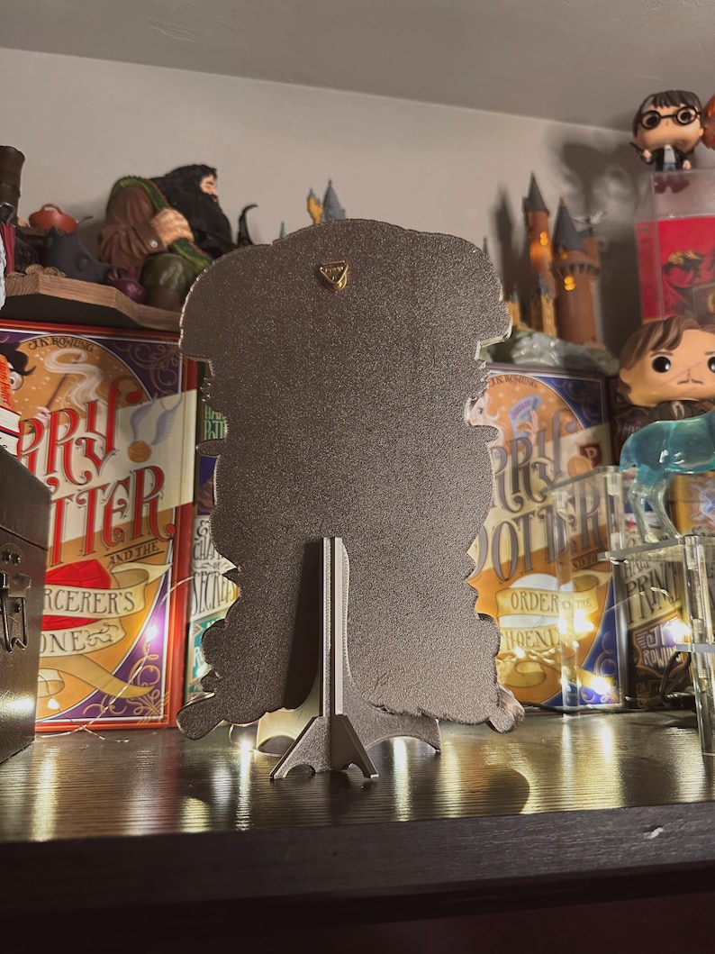 May include: A black metal silhouette of a wizard with a pointed hat and a long beard. The silhouette is on a stand and has a gold accent on the top. The silhouette is in front of a bookshelf with Harry Potter books and figurines.