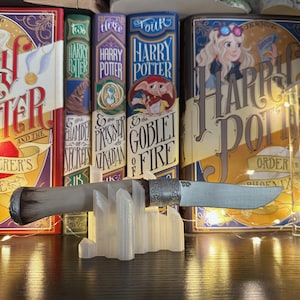 May include: A decorative knife with a silver blade and handle, resting on a white stand. The handle has a dark brown end and silver accents. The knife is displayed in front of a collection of Harry Potter books.