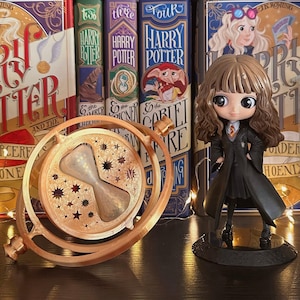 May include: A stylized figurine of a girl with brown hair wearing a black robe and a gold-colored time-turner pendant featuring a central hourglass and rotating rings with star-shaped cutouts, positioned in front of several Harry Potter books.
