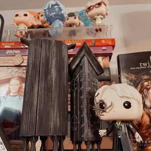 May include: A black and gray Harry Potter Pop figure with a spider on its face, standing in front of two black and gray wooden structures. The structures are tall and narrow, with a pointed top and a base with four legs. The figure is holding its right hand out, as if it is pointing at something.