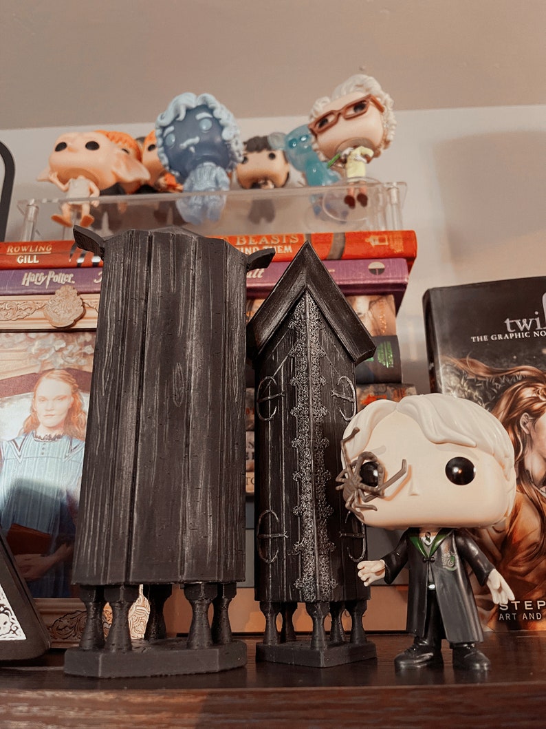 May include: A black and gray Harry Potter Pop figure with a spider on its face, standing in front of two black and gray wooden structures. The structures are tall and narrow, with a pointed top and a base with four legs. The figure is holding its right hand out, as if it is pointing at something.