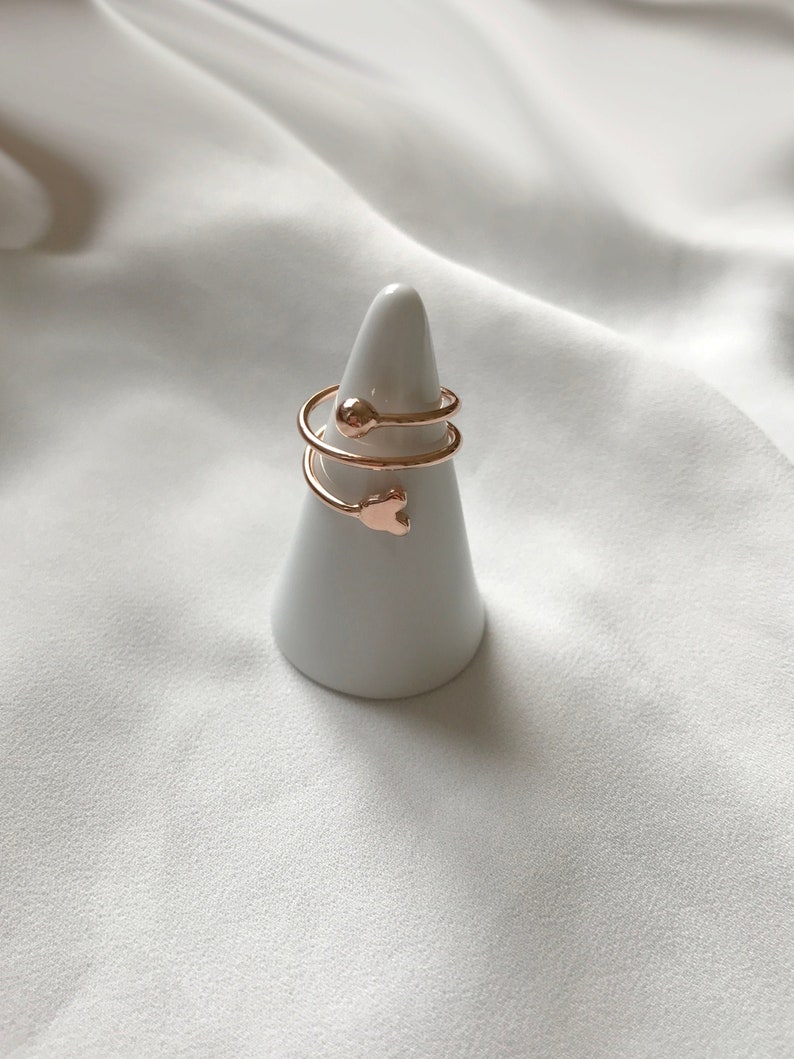 K&ouml;nnte beinhalten: Ein Set aus drei goldfarbenen Ringen mit einem Herz- und einem Kugeldesign. Die Ringe werden auf einem wei&szlig;en, kegelf&ouml;rmigen Ringst&auml;nder pr&auml;sentiert.