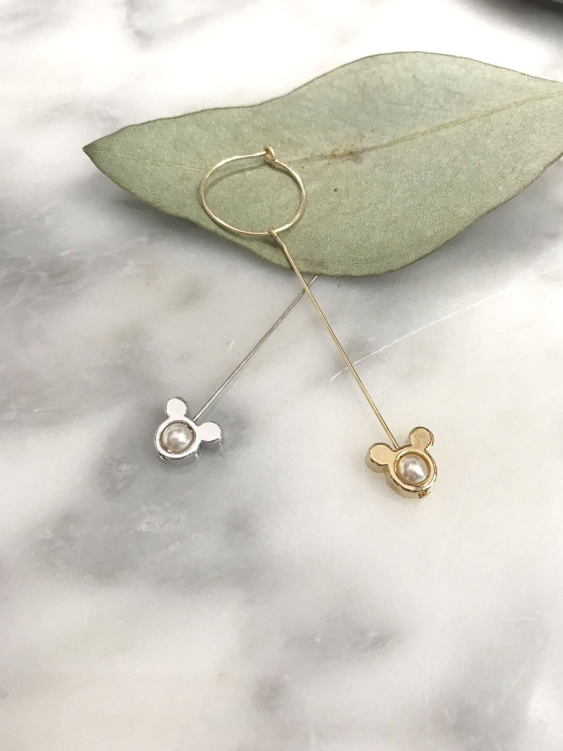 May include: A pair of dangle earrings with a pearl and a Mickey Mouse head design. One earring is gold and the other is silver.