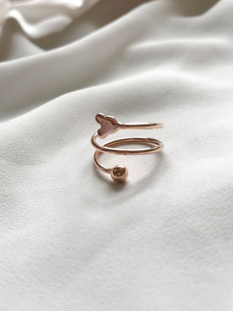 K&ouml;nnte beinhalten: Ein Ring aus Ros&eacute;gold mit einem herzf&ouml;rmigen Design und einer kleinen Kugel am Ende des Bandes.