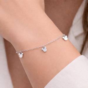 May include: A delicate silver bracelet featuring three small, round charms with a mouse-ear design. The bracelet is worn on a wrist, and the charms dangle from a fine chain. The bracelet is a simple, elegant piece of jewelry.