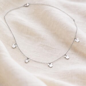 May include: A delicate silver necklace featuring small, round charms with a Mickey Mouse head design. The necklace is displayed on a white, textured fabric, highlighting the jewelry's simple elegance and playful theme.