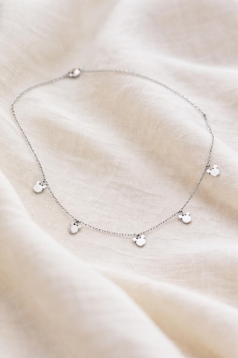 May include: A delicate silver necklace featuring small, round charms with a Mickey Mouse head design. The necklace is displayed on a white, textured fabric, highlighting the jewelry's simple elegance and playful theme.
