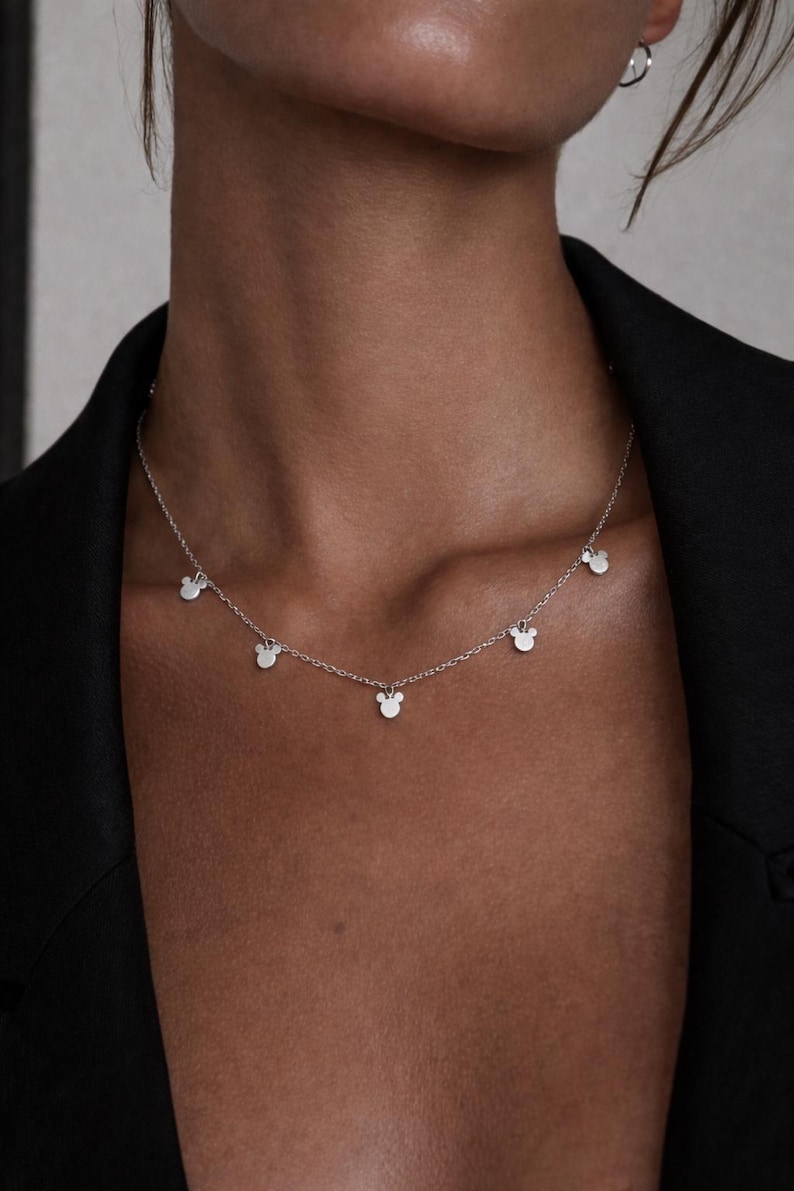 May include: Silver necklace featuring multiple small, stylized mouse head charms. The delicate chain is worn around the neck, with the charms evenly spaced. The necklace is a simple yet elegant piece of jewelry.