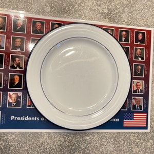 May include: A white dinner plate with a blue rim sits on top of a table runner featuring a grid of portraits of U.S. presidents. The table runner has the text "Presidents of" and "America" printed on it. The bottom of the table runner features a small American flag.