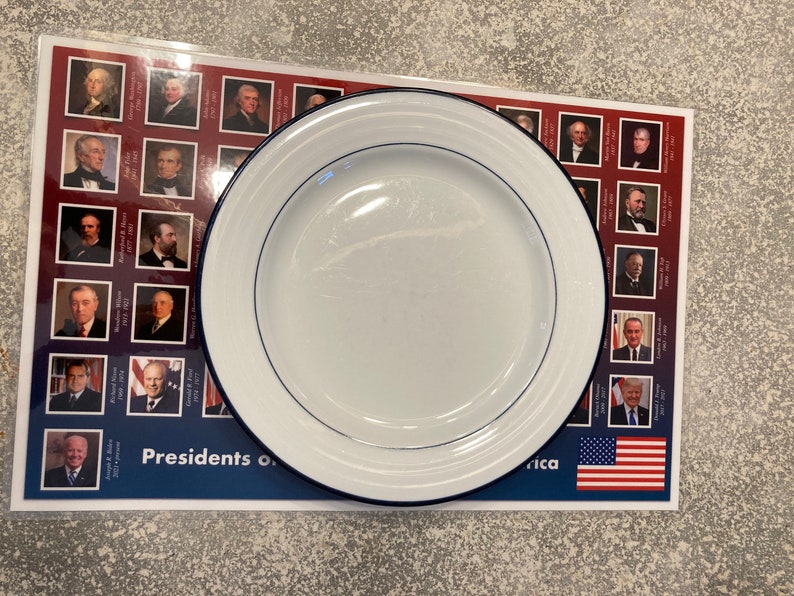 May include: A white dinner plate with a blue rim sits on top of a table runner featuring a grid of portraits of U.S. presidents. The table runner has the text "Presidents of" and "America" printed on it. The bottom of the table runner features a small American flag.