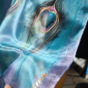 Puede incluir: Un pañuelo de seda en tonos turquesa y azul con un motivo de pluma de pavo real pintado a mano con detalles en morado y dorado. La tela tiene un brillo suave y reflectante e incluye una firma escrita en tinta dorada en el borde inferior.