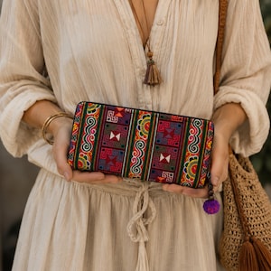 May include: A rectangular wallet featuring a vibrant, multi-coloured embroidered fabric with intricate geometric patterns in red, green, yellow, and purple. The wallet has a black zip closure and a small purple pom-pom attached to the zip pull.