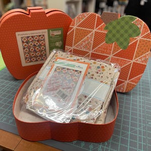May include: Two orange pumpkin-shaped boxes. The larger box has a patchwork design with green and orange fabric. The smaller box contains a fabric bundle with a floral pattern and a label that reads "Embroidery Sampler".