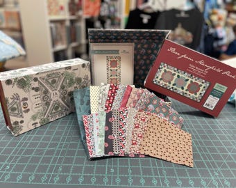 May include: A quilting kit for a table runner featuring a floral design with red, green, and white fabrics. The kit includes a variety of pre-cut fabric squares and a pattern guide. The kit is titled "Flora from Mansfield Park Table Runner Kit".