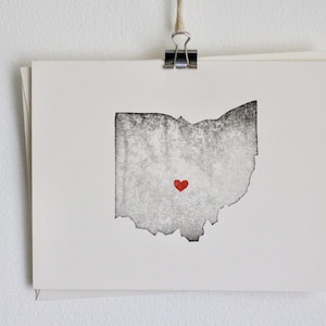 Op de afbeelding: Een zwart-witte print van de staat Ohio met een rood hart in het midden.