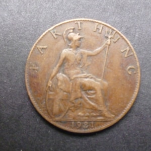 Könnte beinhalten: Eine kupferfarbene britische Münze mit der Aufschrift "ARTHUR HING" und dem Jahr "1921" auf der Rückseite. Die Münze zeigt eine sitzende Figur von Britannia, die einen Dreizack hält.