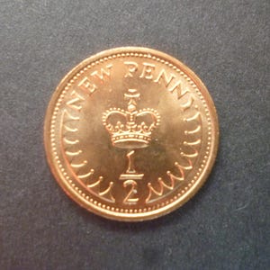 Può includere: Una moneta di rame da mezzo New Penny. La moneta presenta la scritta "NEW PENNY", una corona e la frazione "1/2". La moneta è posta su uno sfondo grigio scuro.