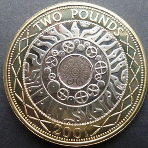 Puede incluir: Moneda de dos libras esterlinas del año 2001. La moneda presenta un diseño dorado y plateado con la inscripción "TWO POUNDS" y el año "2001". El diseño central incluye engranajes y un patrón solar.