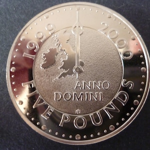 May include: A silver five pound coin from the United Kingdom, commemorating the year 2000. The coin features a map of the British Isles with a clock hand pointing to the year 2000. The inscription reads "1999 Anno Domini 2000 Five Pounds".