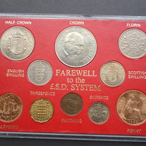 Op de afbeelding: Een verzameling Britse munten, waaronder een Half Crown, Crown, Florin, English Shilling, Scottish Shilling, Halfpenny, Threepence, Farthing, Sixpence en Penny. De munten worden gepresenteerd op een rode achtergrond met de tekst "FAREWELL to the £.S.D. SYSTEM."
