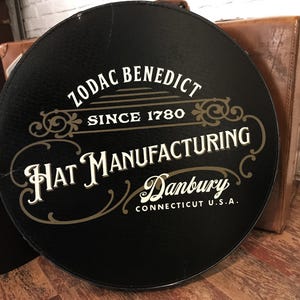 Op de afbeelding: Zwarte hoedendoos met gouden opschrift "Zodiac Benedict Since 1780 Hat Manufacturing Danbury Connecticut U.S.A."