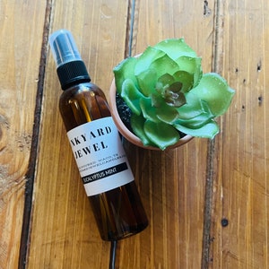 May include: A brown glass bottle with a black spray nozzle and a white label that reads "Backyard Jewel Hand Poured Waco, TX BackyardJewelCandles.com Eucalyptus Mint".