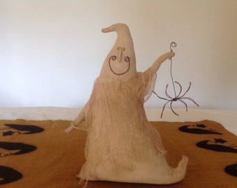 May include: A white fabric ghost doll with a stitched-on smile and eyes, holding a wire spider. The doll is sitting on a brown fabric surface with black fabric spider silhouettes.
