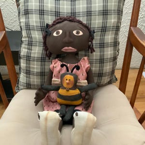 May include: A handmade doll with dark skin, wearing a pink dress with a bee design. The doll is holding a stuffed bee toy. The doll has brown hair and is sitting on a cream-colored chair with a plaid pillow in the background.