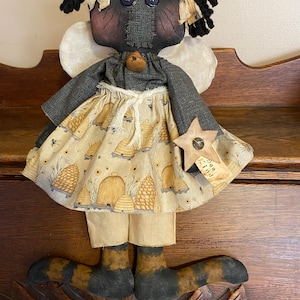May include: A black rag doll with a bee-themed dress and wings. The doll is holding a star-shaped tag that says "Bee Kind".