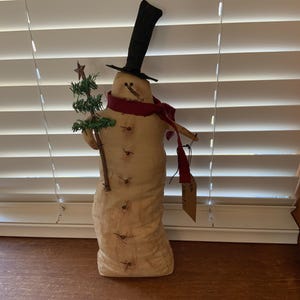 May include: A fabric snowman figurine with a black top hat, red scarf, and a small green Christmas tree. The snowman has button details and a tag hanging from its arm. The figurine is standing on a wooden surface.