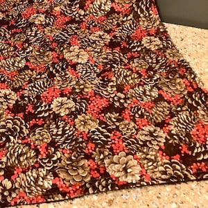 May include: A brown fabric placemat with a repeating pattern of gold pine cones and red berries.