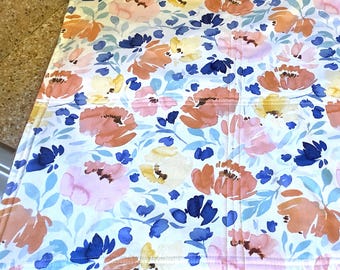 May include: A white kitchen dish towel with a floral pattern in shades of pink, orange, blue, and yellow. The towel has a brown border.