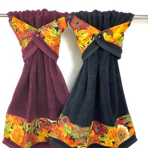 May include: Two decorative hand towels, one burgundy and one black, hang from a metal bar. Each towel features a floral pattern with sunflowers, autumn leaves, and a coordinating border. The towels are designed with a decorative top and a button closure.